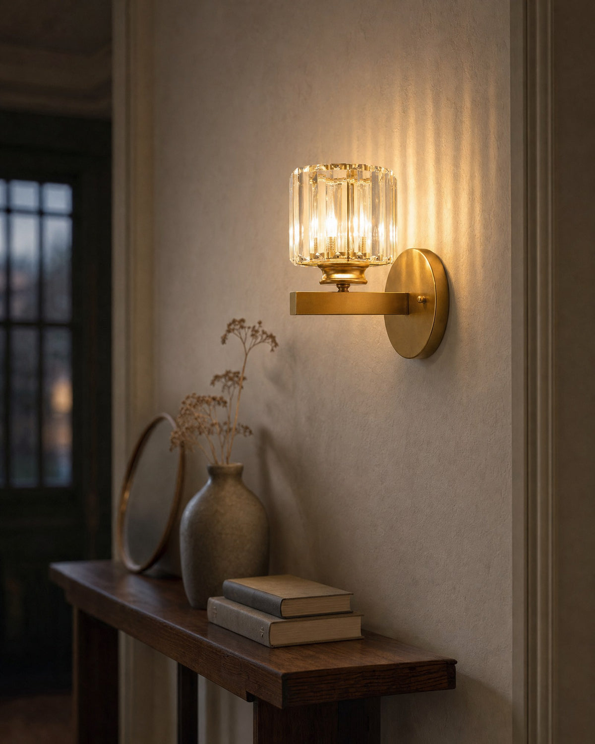 Satin gold crystal cylinder Art Deco wall light glowing at dusk above an entryway console, prismatic light on the plaster wall