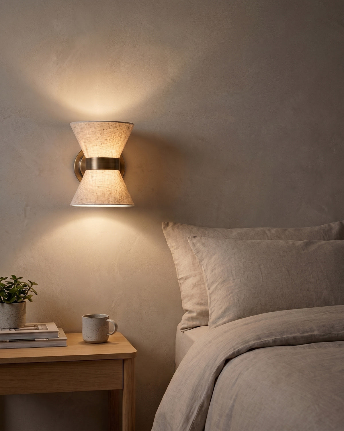 Halston linen and bronze wall light glowing warm beside a bed in a bright bedroom