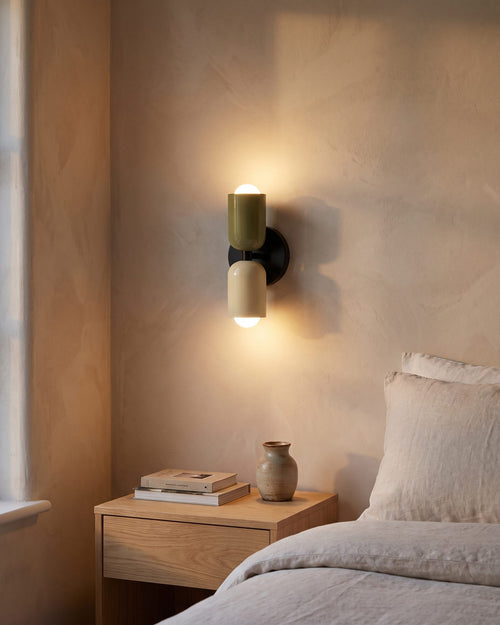 Green and cream up-and-down wall light glowing warm beside a bed in a bright bedroom