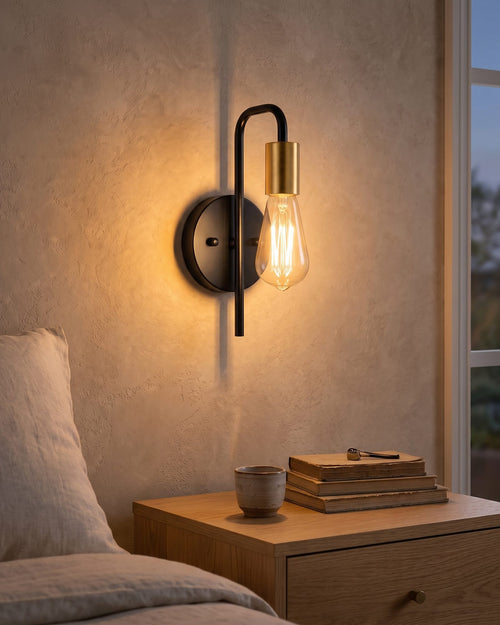 Black and gold bare bulb wall light glowing warm above a bedside table in a bright bedroom