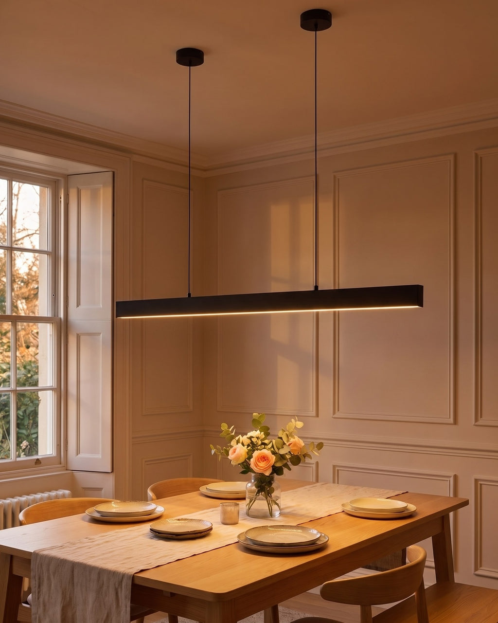 Thala linear LED pendant in matte black glowing over a laid dining table at dusk