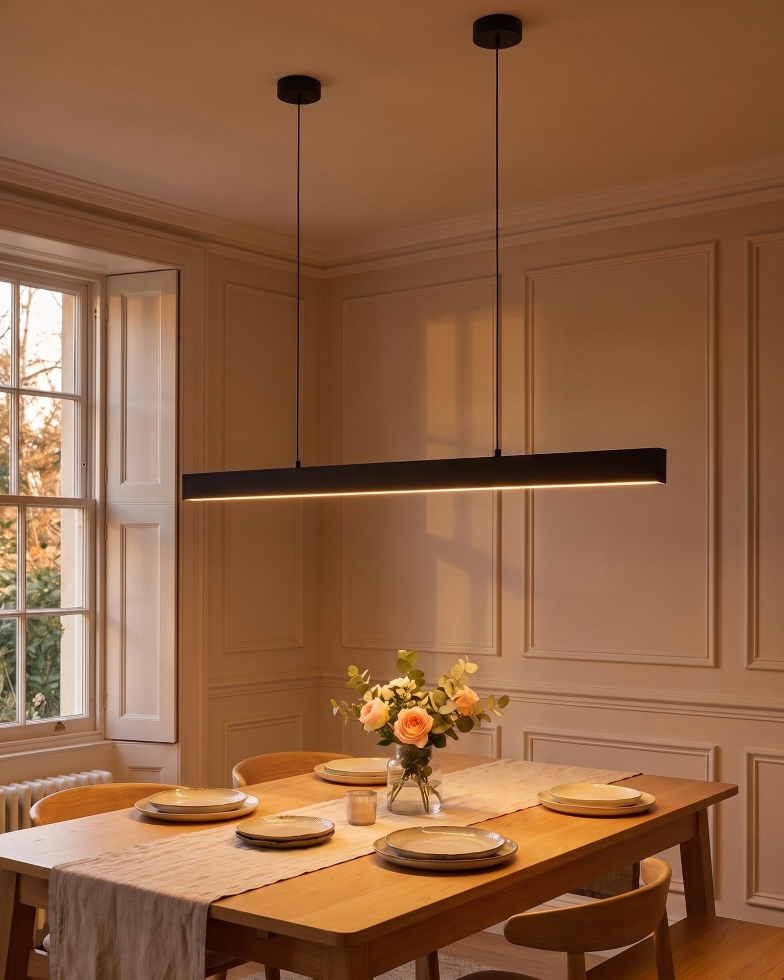 Thala linear LED pendant in matte black glowing over a laid dining table at dusk